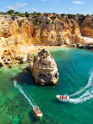 Tourists on boats exploring coastal caves near a sandy beach in Algarve, Portugal.