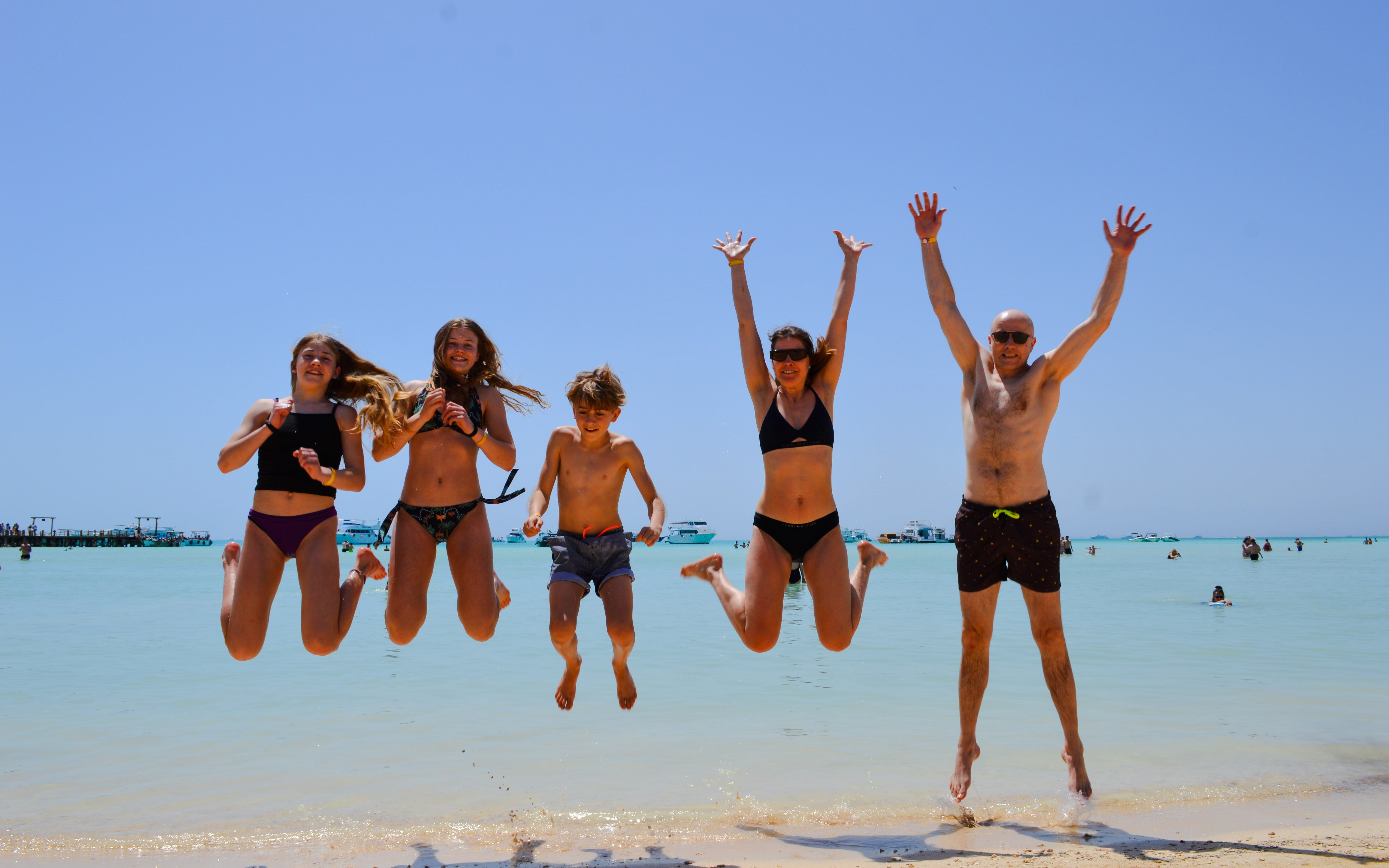 Group jumping on beach during Giftun 3 Islands Tour, clear water and boats in background.
