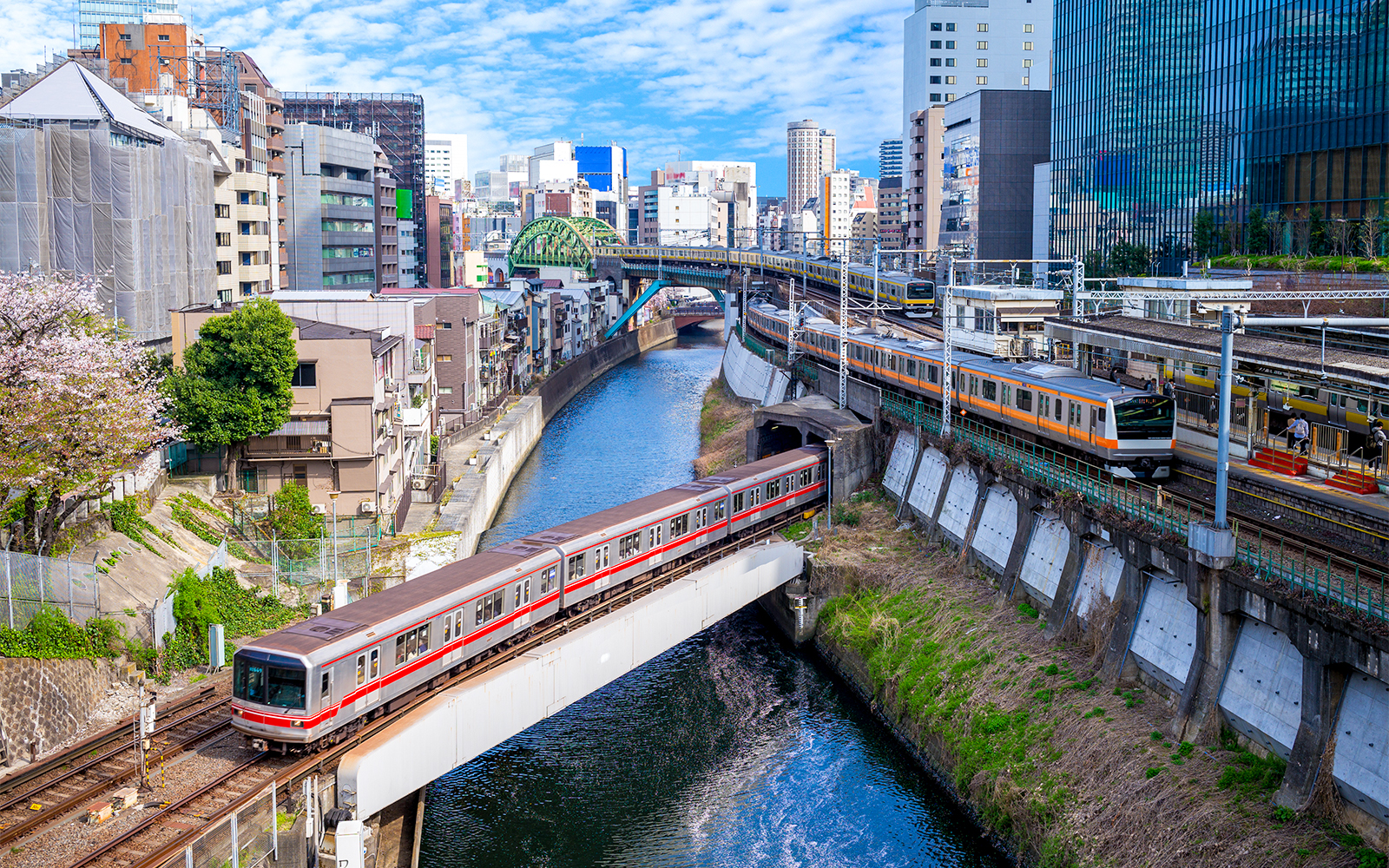 Train crossing a bridge over a canal in Tokyo with cityscape in the background.