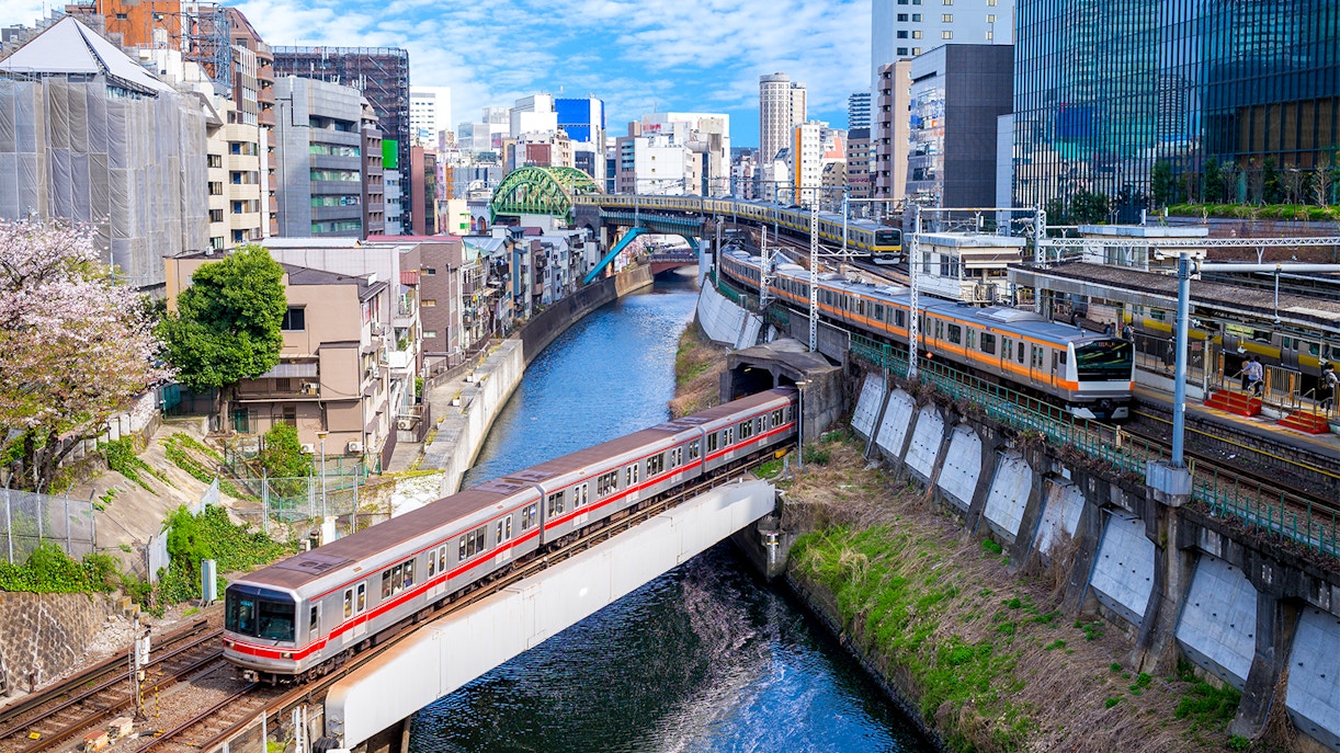 Train crossing a bridge over a canal in Tokyo with cityscape in the background.