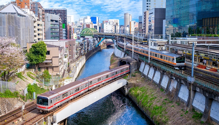 Train crossing a bridge over a canal in Tokyo with cityscape in the background.