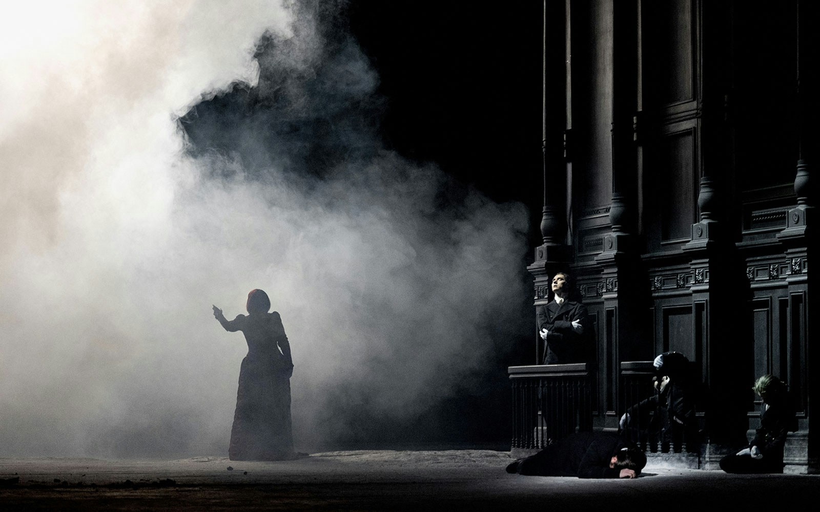 Opera scene from Salome with a silhouetted figure in smoke and performers by a dark structure.