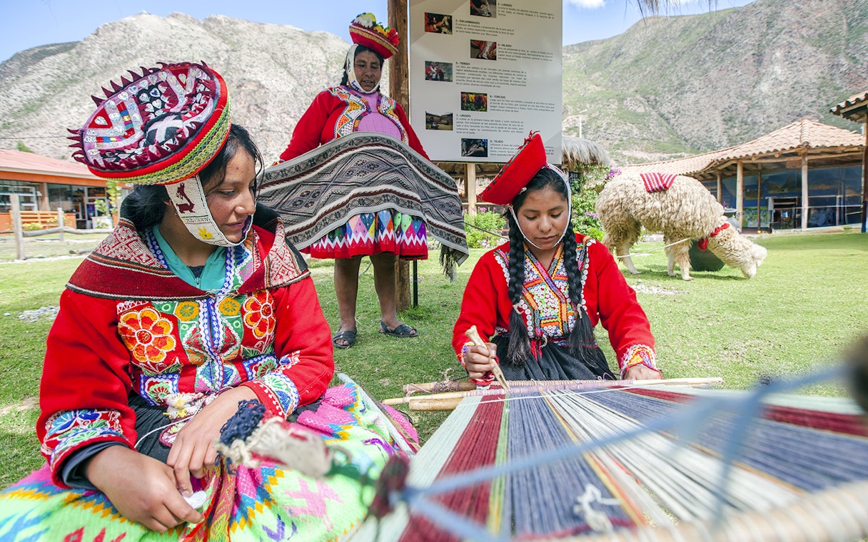 Peruvian women in traditional attire weaving outdoors with mountains in the background.