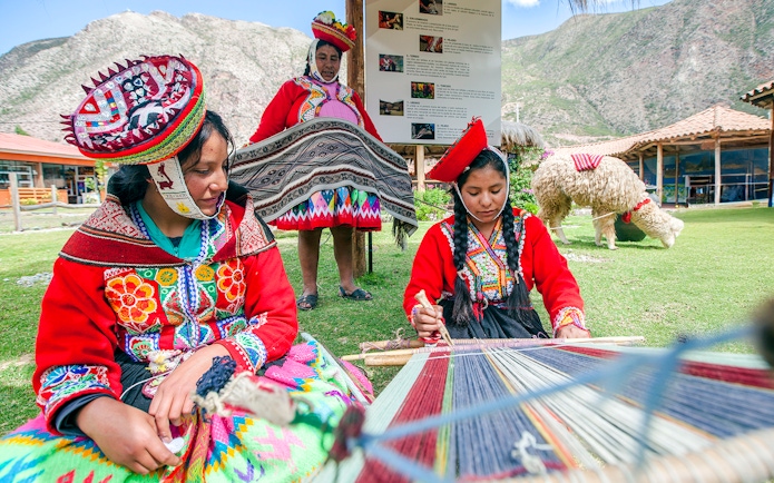 Peruvian women in traditional attire weaving outdoors with mountains in the background.