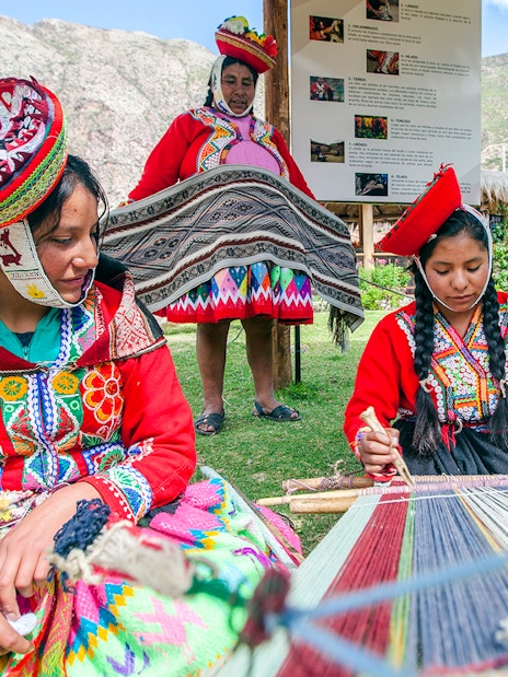Peruvian women in traditional attire weaving outdoors with mountains in the background.