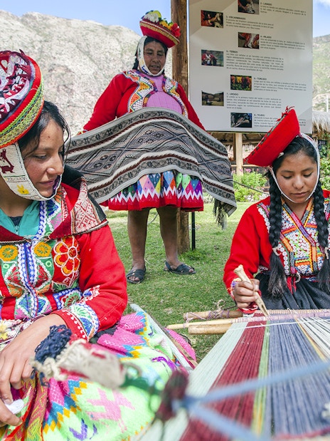 Peruvian women in traditional attire weaving outdoors with mountains in the background.