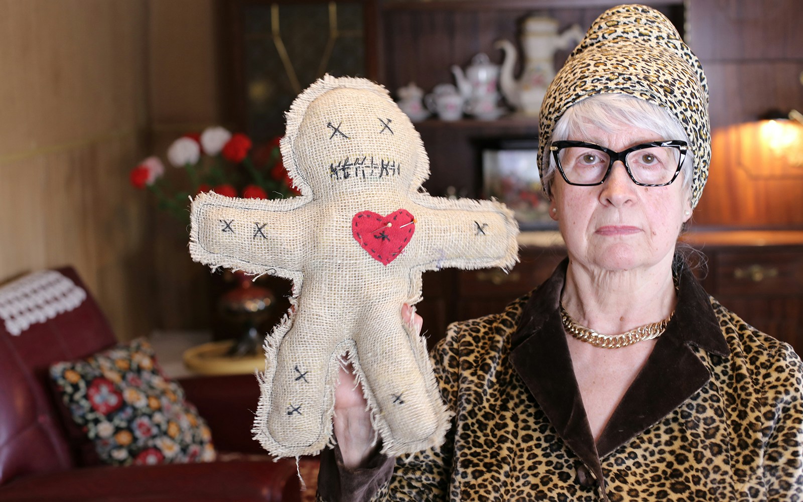Senior woman holding a burlap voodoo doll with a red heart in a vintage room.