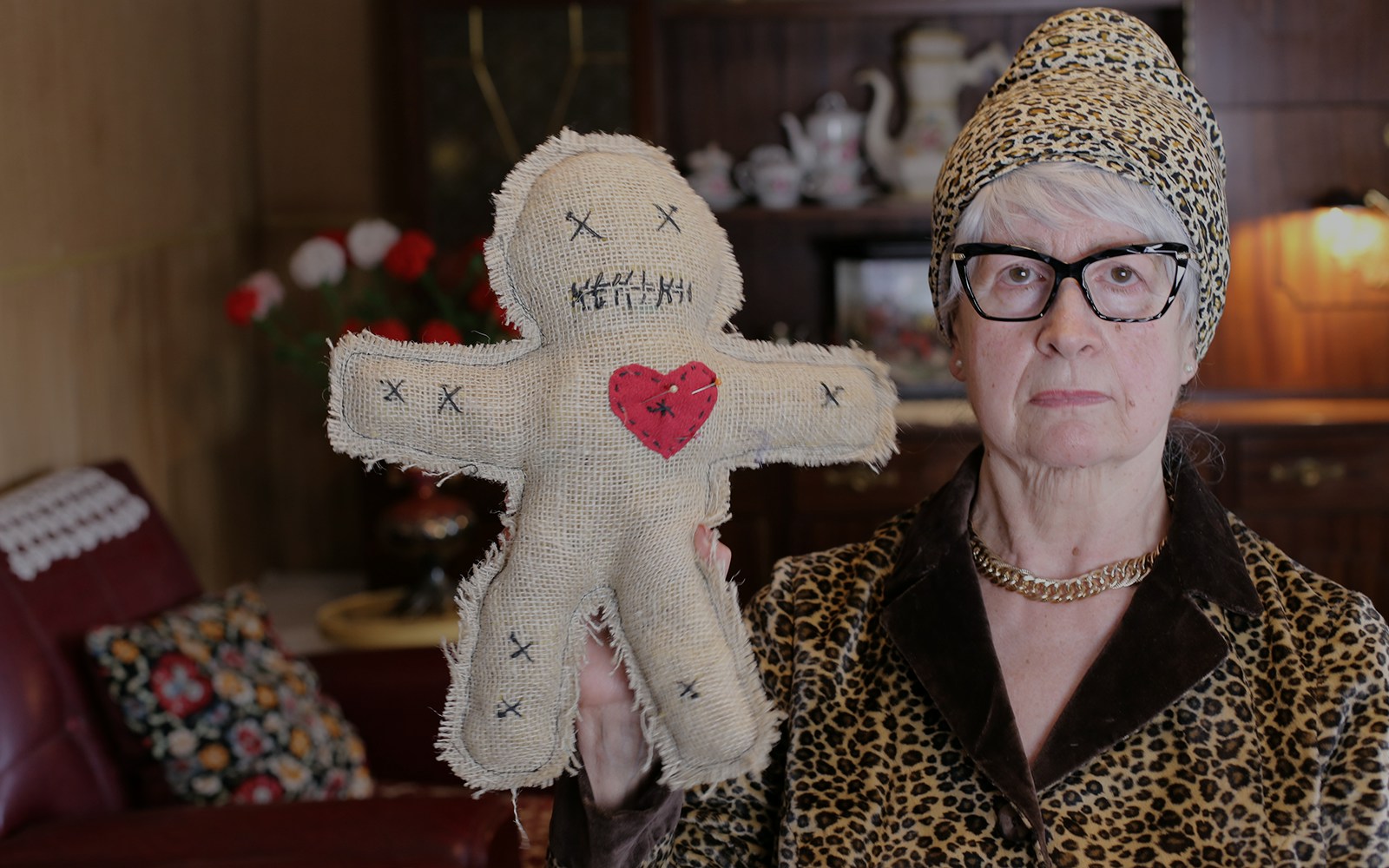 Senior woman holding a burlap voodoo doll with a red heart in a vintage room.
