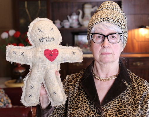 Senior woman holding a burlap voodoo doll with a red heart in a vintage room.