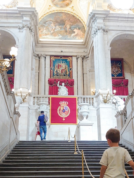 Grand staircase inside the Royal Palace of Madrid with ornate chandeliers and royal emblems.