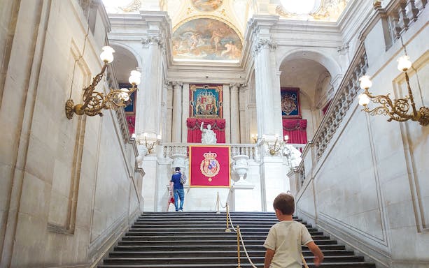 Grand staircase inside the Royal Palace of Madrid with ornate chandeliers and royal emblems.