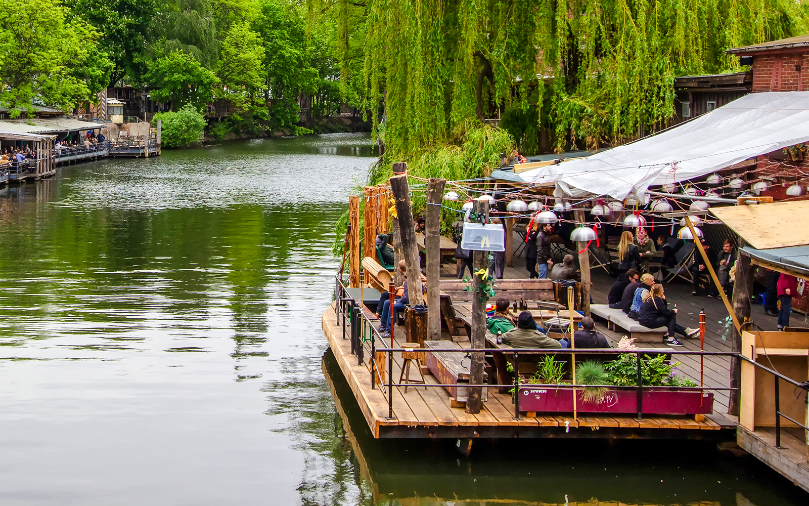 Cafe terrace along the Spree River in Berlin with people enjoying drinks.