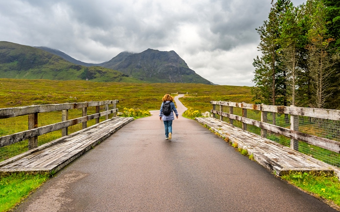 Female hiker walking on a bridge in Glencoe, Scotland, with mountains in the background.