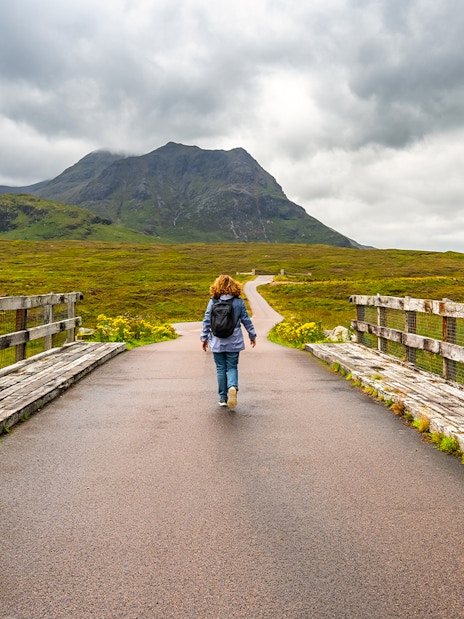 Female hiker walking on a bridge in Glencoe, Scotland, with mountains in the background.