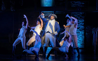 Hamilton cast performing on West End stage, actors in period costumes.