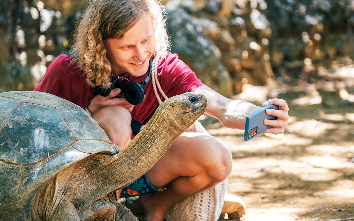 Tourist taking a selfie with an Aldabra Tortoise in a natural setting.