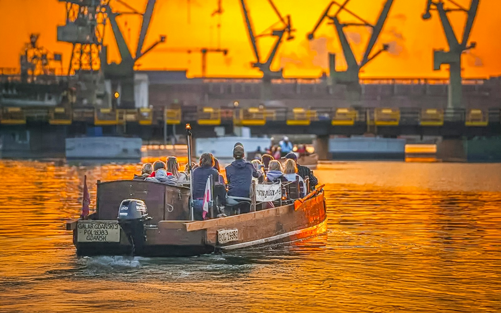 Guests on a historical boat cruise at sunset, viewing Gdansk Shipyard cranes.