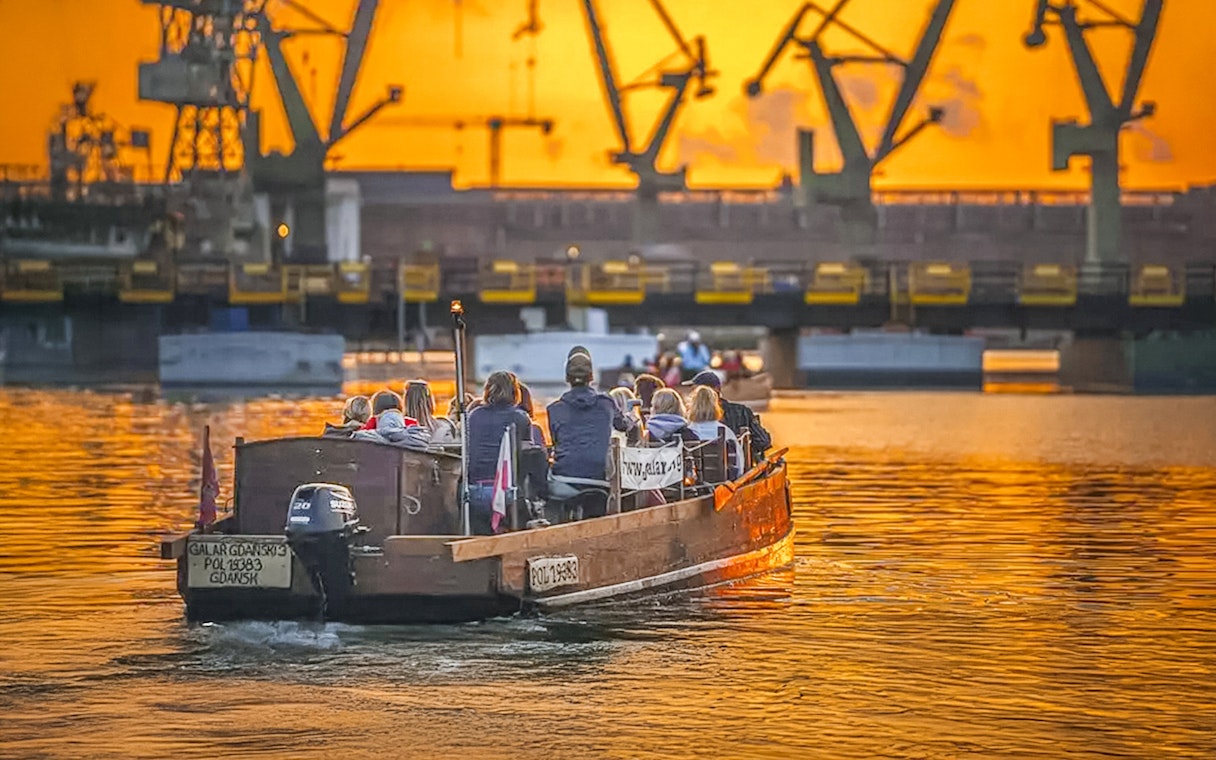 Guests on a historical boat cruise at sunset, viewing Gdansk Shipyard cranes.
