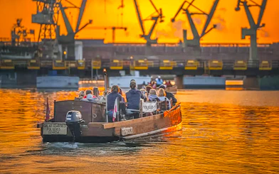 Guests on a historical boat cruise at sunset, viewing Gdansk Shipyard cranes.