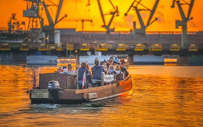 Guests on a historical boat cruise at sunset, viewing Gdansk Shipyard cranes.