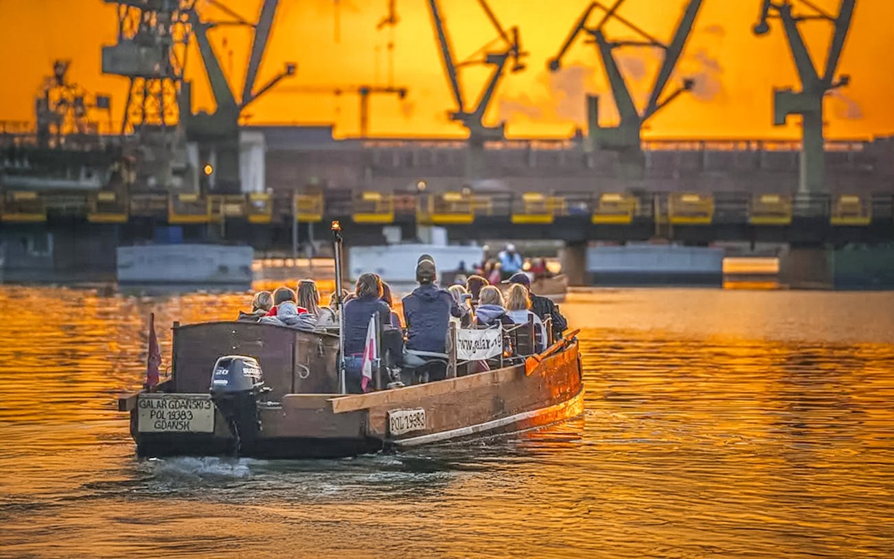 Guests on a historical boat cruise at sunset, viewing Gdansk Shipyard cranes.