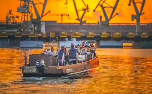 Guests on a historical boat cruise at sunset, viewing Gdansk Shipyard cranes.