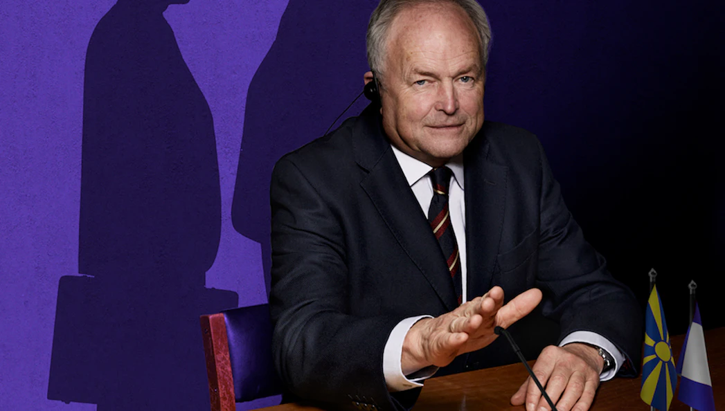 Man in suit at conference table with flags, shadow on purple wall.