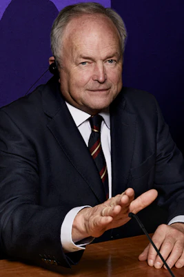 Man in suit at conference table with flags, shadow on purple wall.
