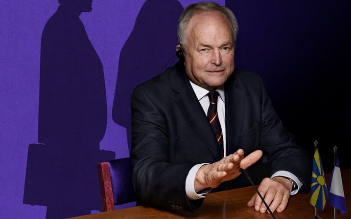 Man in suit at conference table with flags, shadow on purple wall.