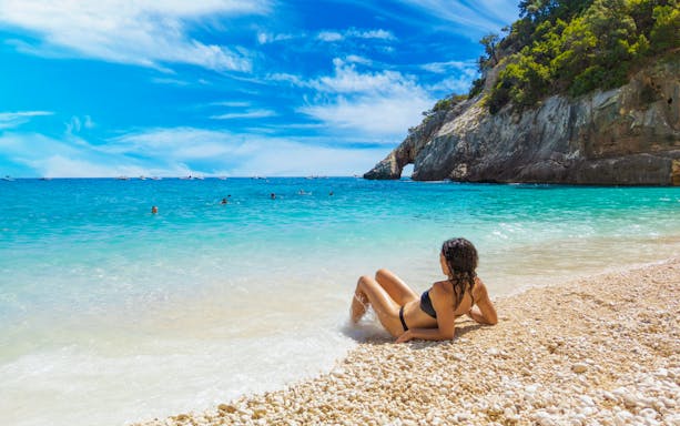 Person relaxing on a pebble beach by the turquoise sea in Gulf of Orosei, Sardinia, Italy.