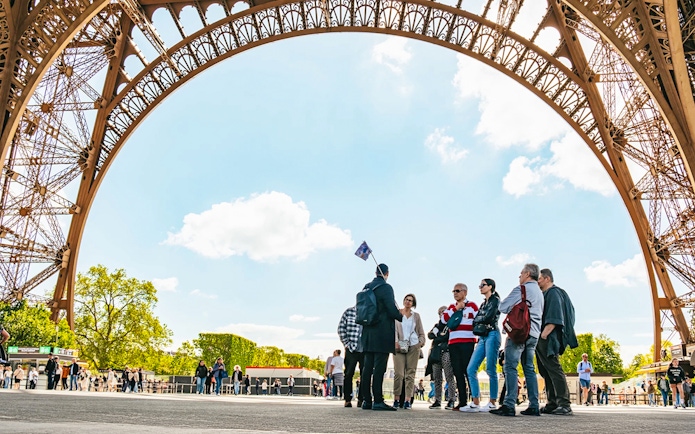 Tour group under Eiffel Tower arch, Paris, preparing for guided elevator ascent.