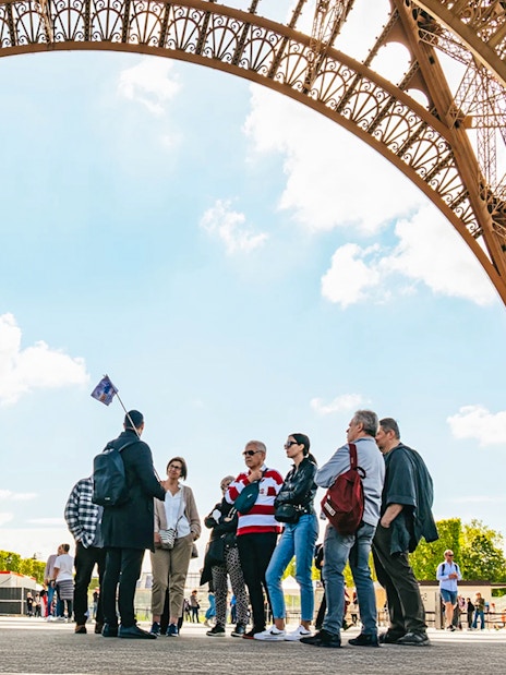 Tour group under Eiffel Tower arch, Paris, preparing for guided elevator ascent.