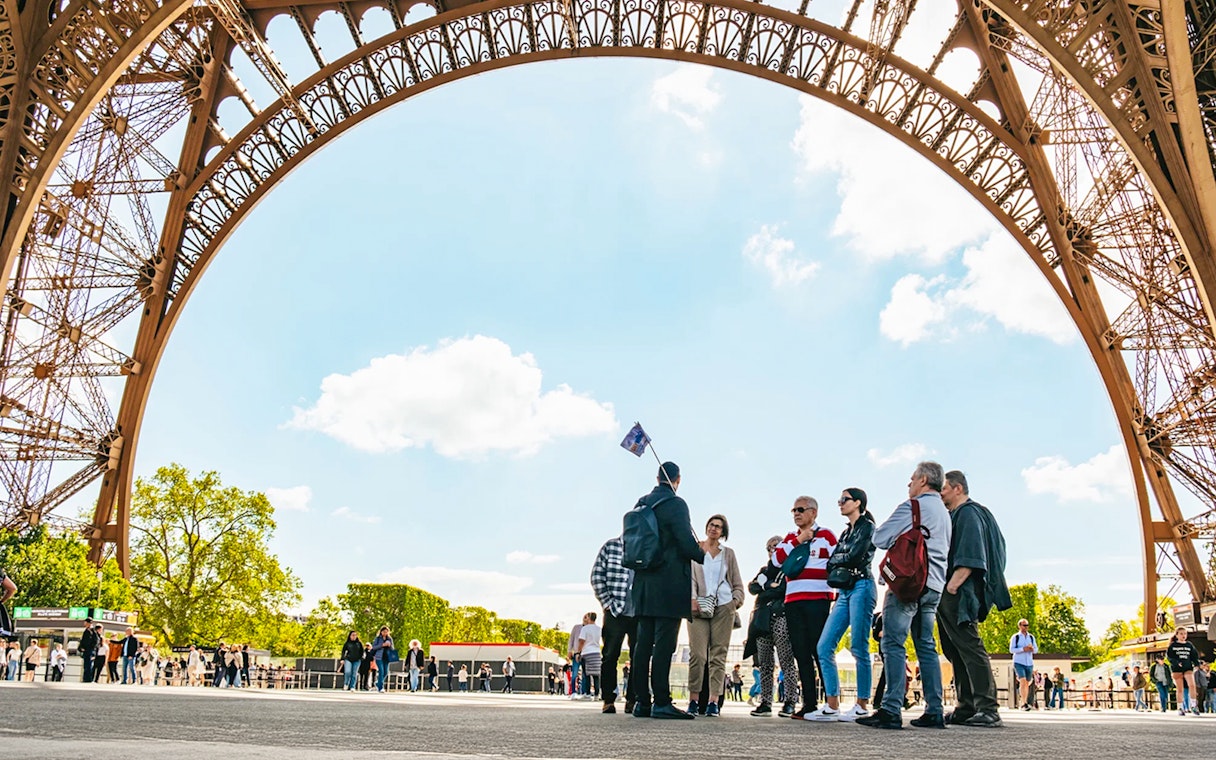 Tour group under Eiffel Tower arch, Paris, preparing for guided elevator ascent.