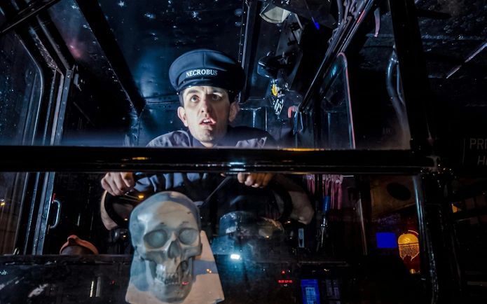 Bus driver in uniform steering the London Ghost Bus with a skull decoration on the dashboard.