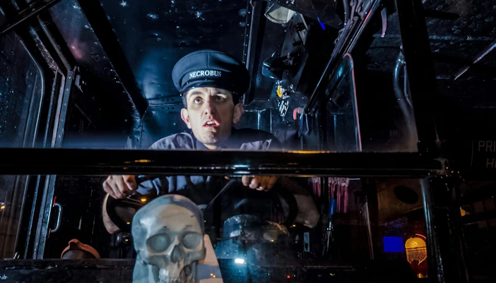 Bus driver in uniform steering the London Ghost Bus with a skull decoration on the dashboard.