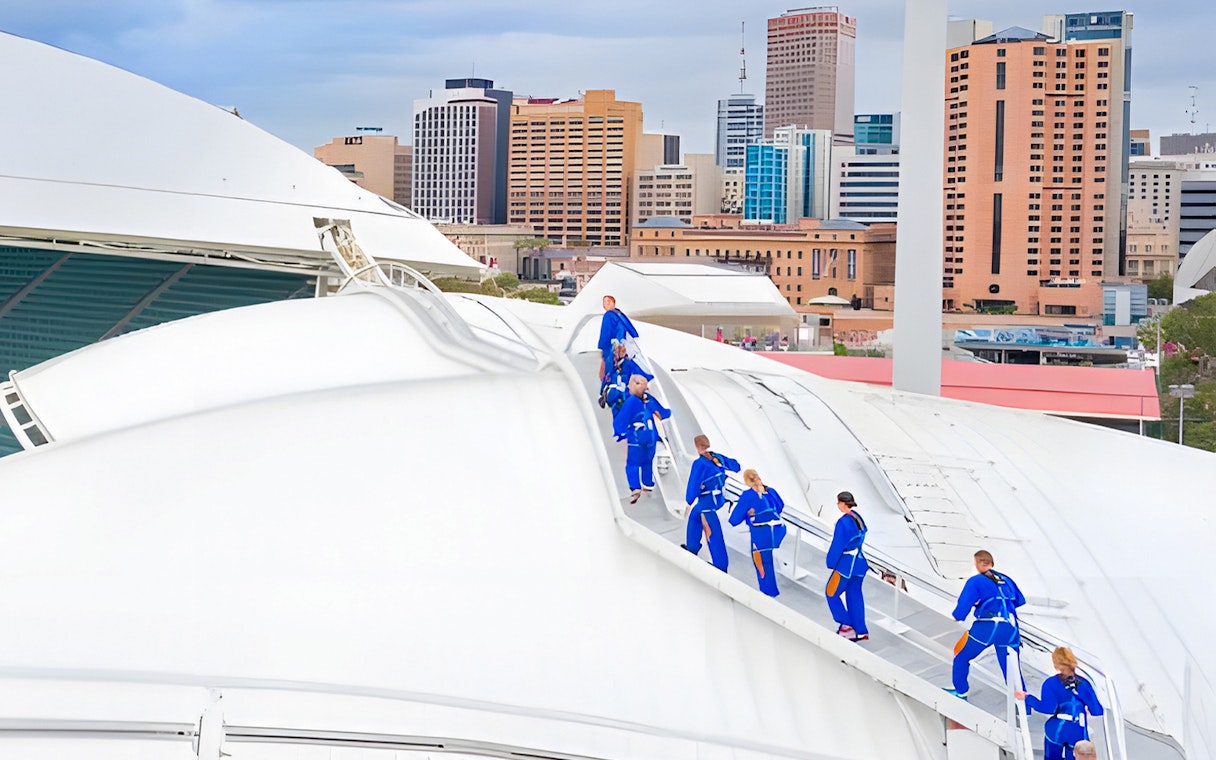 Group climbing Adelaide Oval rooftop with city skyline in background.