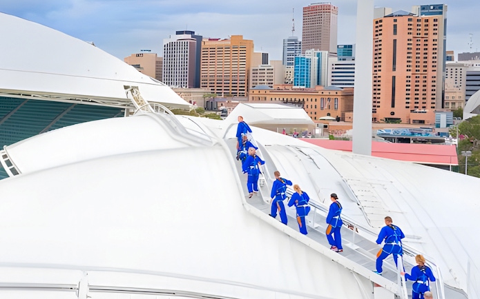 Group climbing Adelaide Oval rooftop with city skyline in background.
