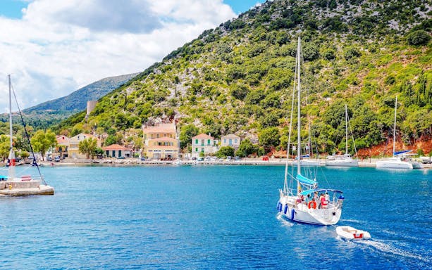 Sailboat approaching Ithaca Island harbor, Greece, with colorful buildings and lush hills in the background.