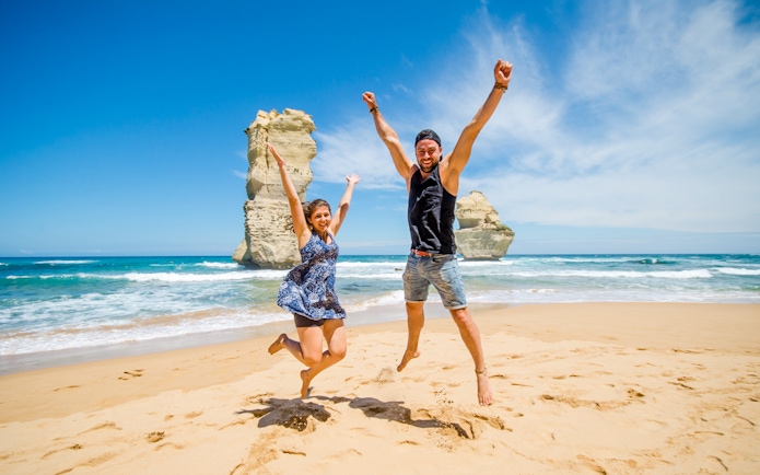 Tourists jumping on the beach at Loch Ard Gorge, Great Ocean Road, with rock formations in the background.