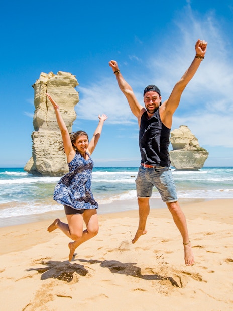 Tourists jumping on the beach at Loch Ard Gorge, Great Ocean Road, with rock formations in the background.