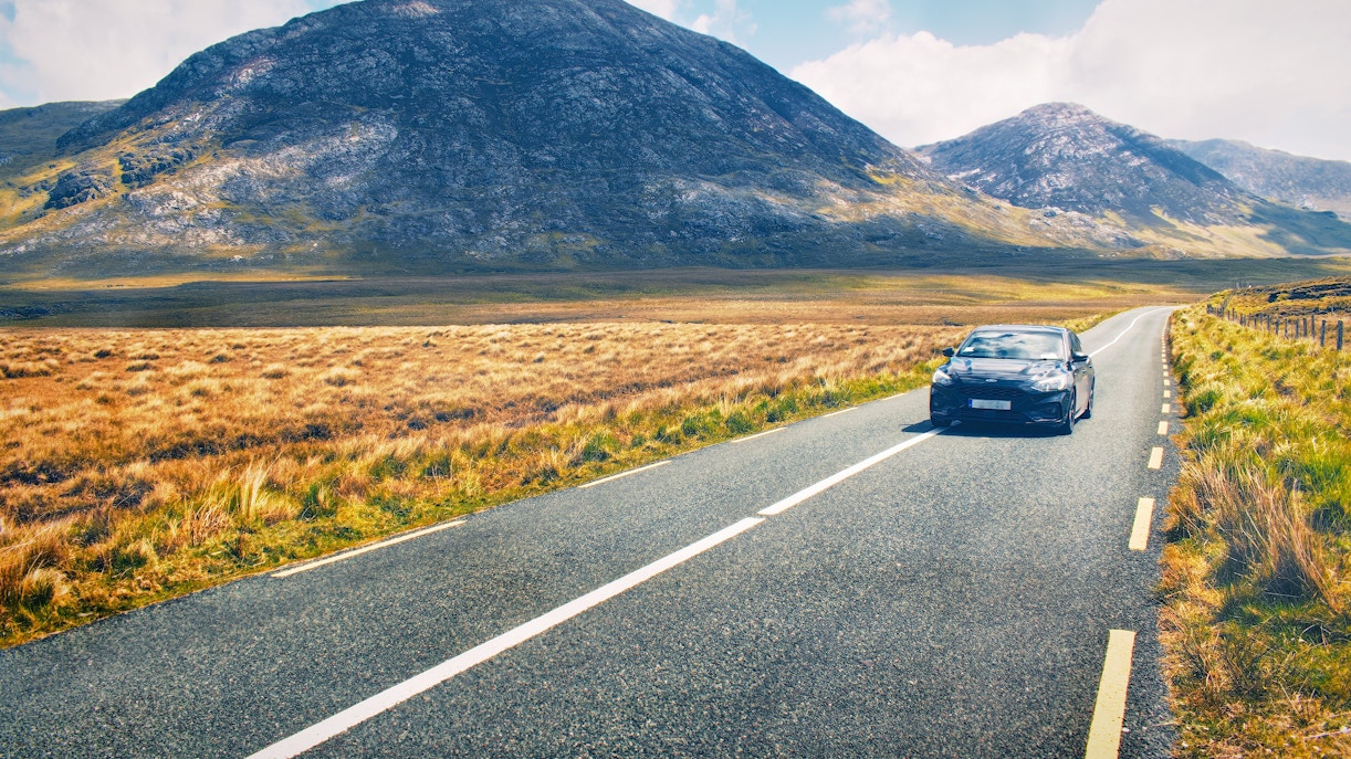 Car driving on a scenic road through the Irish countryside with mountains in the background.