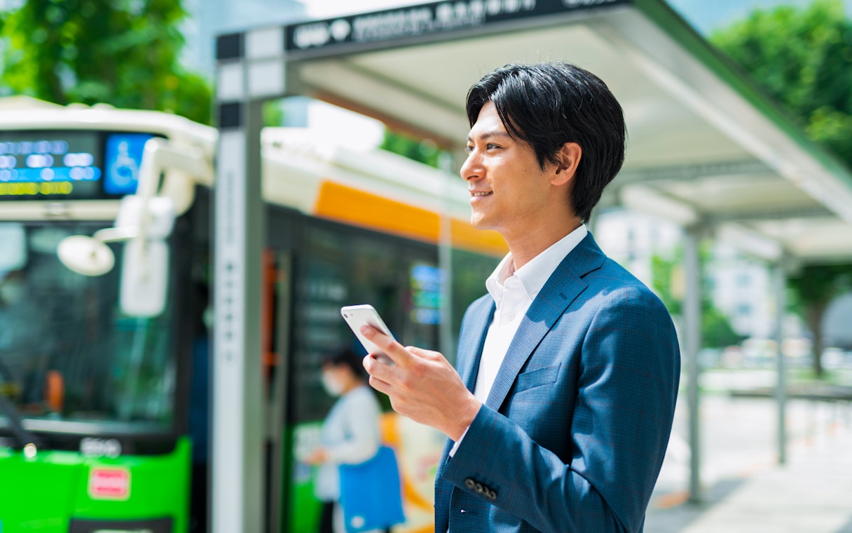 Man using smartphone at Tokyo bus stop with Greater Tokyo Pass.