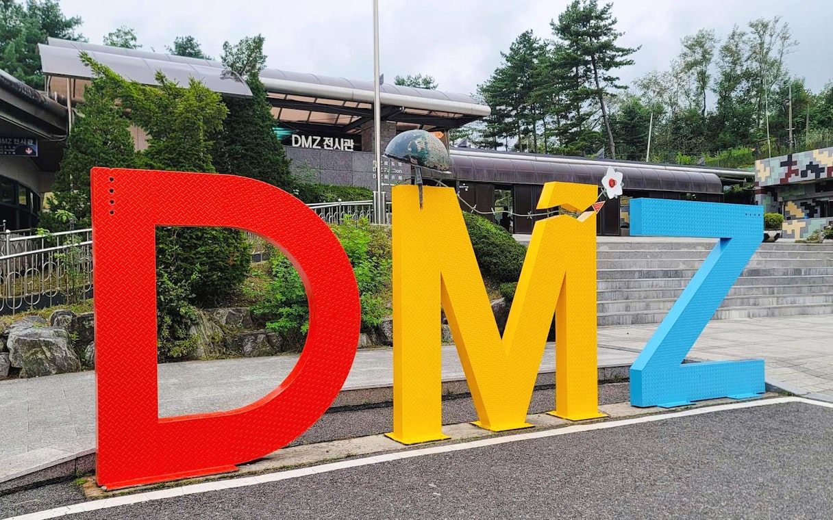 DMZ sign at entrance of exhibition hall in Korea, surrounded by trees and steps.