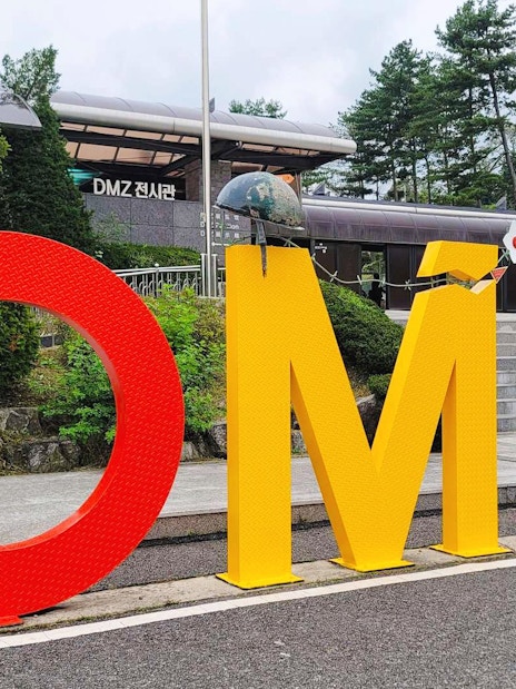 DMZ sign at entrance of exhibition hall in Korea, surrounded by trees and steps.