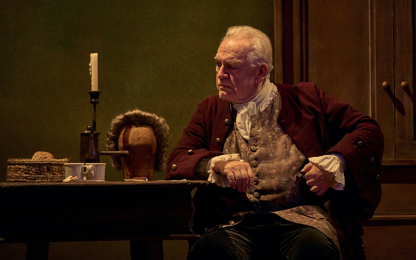 Elderly man in period costume sitting at a table with a candle and wig, related to "The Score.