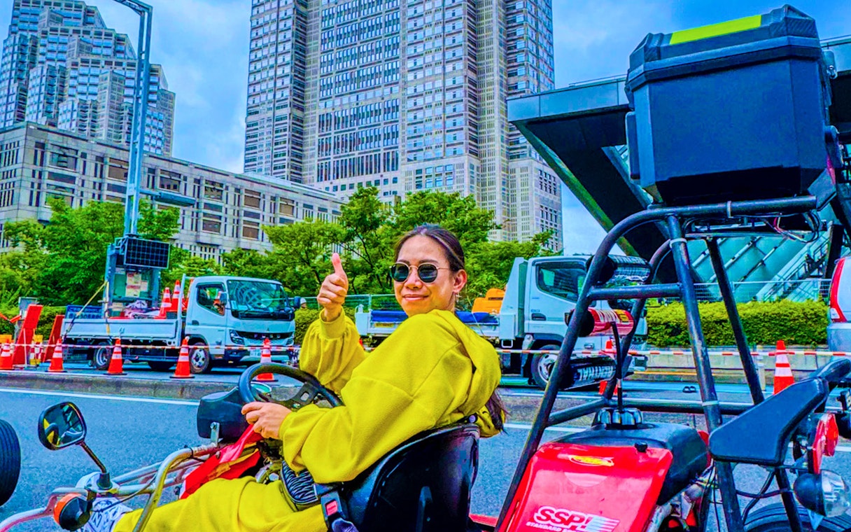 Woman giving thumbs up in go-kart on Shibuya street, Tokyo skyscrapers in background.