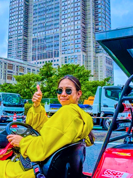 Woman giving thumbs up in go-kart on Shibuya street, Tokyo skyscrapers in background.