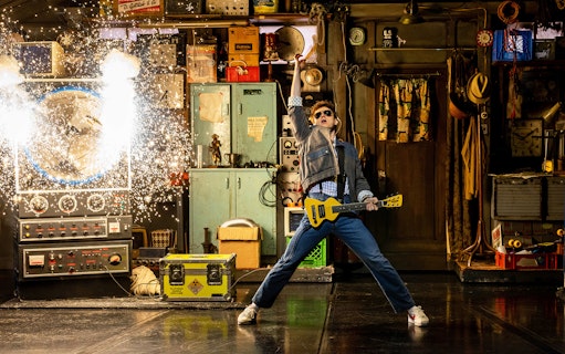 Man in denim jacket with guitar in a workshop, sparks flying from a machine, Back to the Future theme.