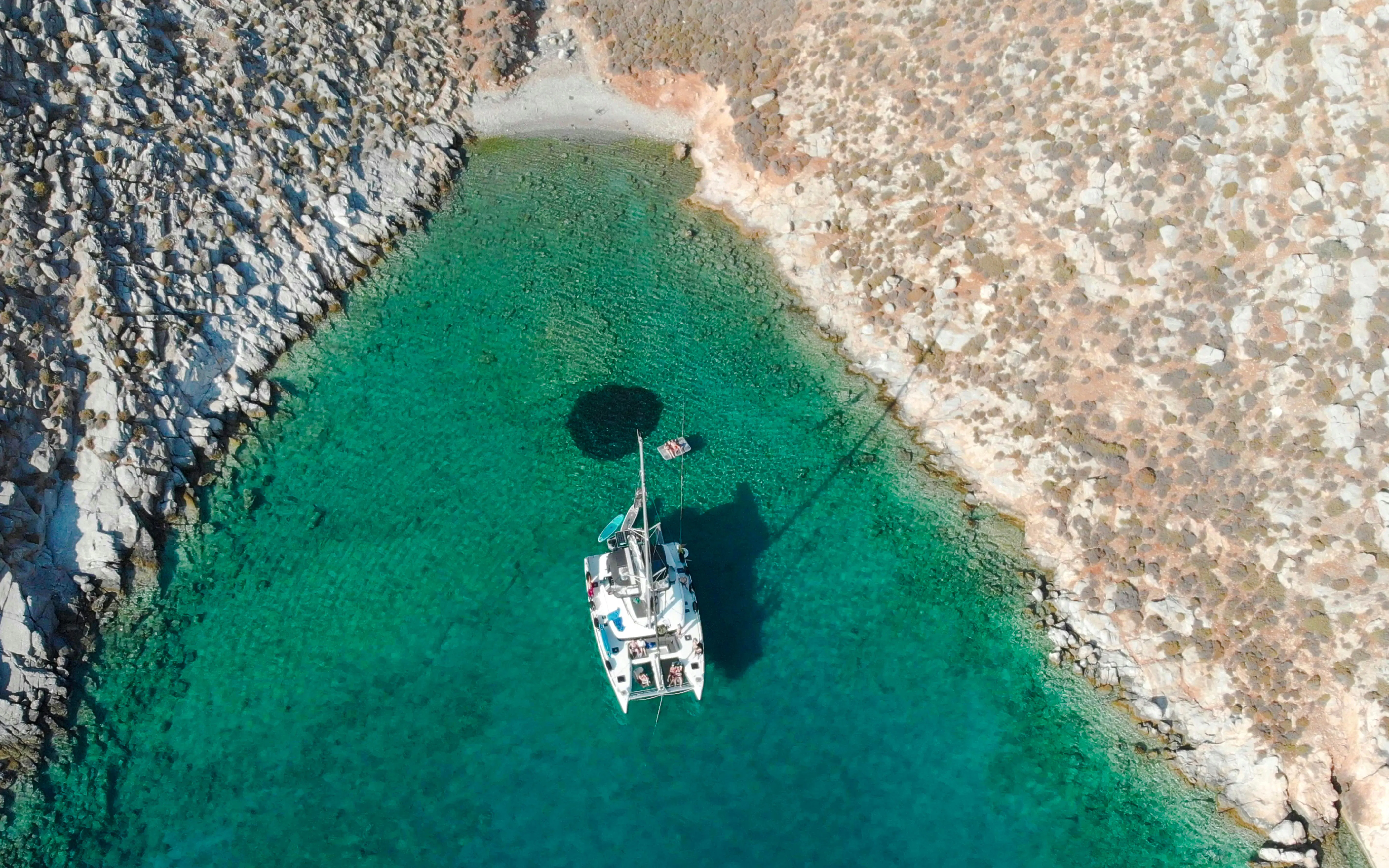 Catamaran anchored in clear waters near rocky Heraklion coastline.