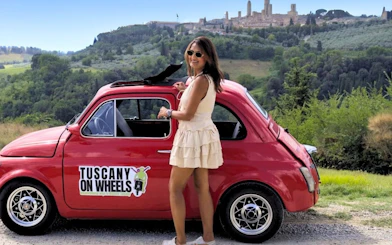 Vintage Fiat 500 parked with scenic view of Tuscany hills and San Gimignano in the background.